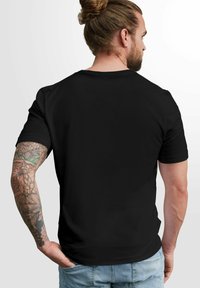 Schwarzes T-Shirt mit kurzen Ärmeln und Rundhalsausschnitt, taillierte Passform und weicher Stoff. Sichtbare Tattoos am Arm verleihen dem Gesamtbild eine zusätzliche Detailtiefe.
