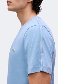 Άνδρας που φοράει ανοιχτό μπλε μπλουζάκι Lacoste με λογότυπο στο στήθος και επώνυμες ρίγες στον ώμο και το μανίκι.
