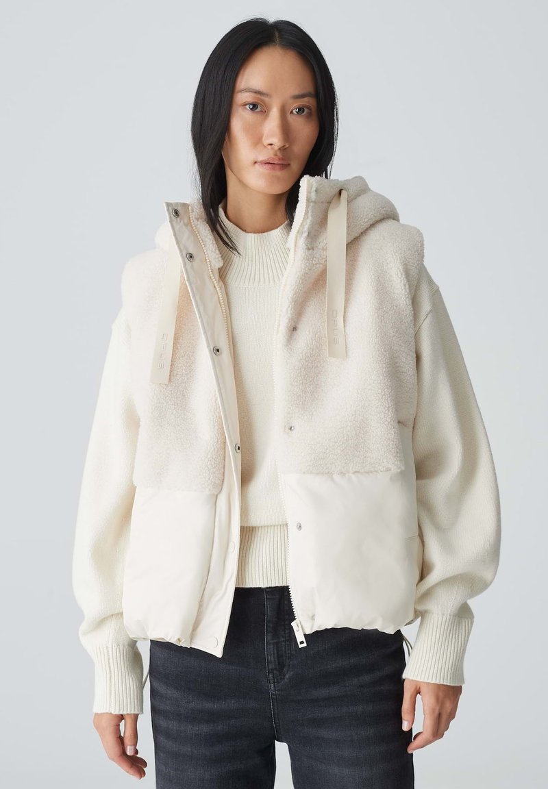 Weißer cropped Blazer mit einem flauschigen oberen Teil, glattem unteren Teil, großer Kapuze und Fronttaschen; getragen über einem cremefarbenen Pullover mit dunklen Jeans.