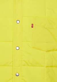 Žltá nafukovacia bunda so prešitými panelmi, striebornými cvokmi a viditeľnou náprsné kapsou s červenou nálepkou Levi's.