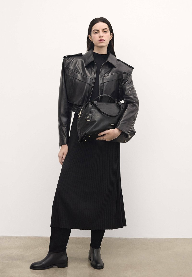 Black leather cropped jacket with shoulder pads, paired with a ribbed black midi dress. The model holds a black leather handbag with gold accents.