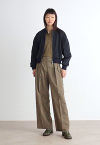 Zwarte bomberjack, olijfgroen geplooid shirt, beige geruite wijde broek, groene schoenen. Soepele texturen en contrasterend kleurenpalet.