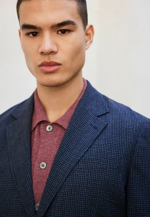 Junger Mann mit kurzen dunklen Haaren, der einen dunkelblauen karierten Blazer über einem bordeauxfarbenen, geknöpften Hemd vor einem einfachen hellen Hintergrund trägt.