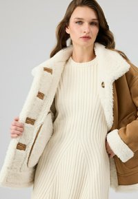 Bruine jas met een witte bontvoering, voorzien van leren accenten en knoopsluitingen. Draagt over een witte ribgebreide jurk met diagonale patronen.