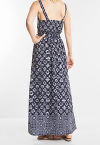 Robe maxi en tissu bleu marine avec motif floral blanc, bretelles ajustables, dos froncé et design d'ourlet blanc contrastant. Sandales plates visibles.