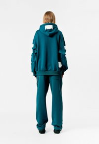Sudadera de gran tamaño en color verde azulado con diseño recortado en las mangas, combinada con pantalones de chándal a juego. Cuenta con un bolsillo tipo canguro y un bolsillo lateral con cremallera.