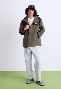 Olijfgroene buitenjas met zwarte accenten, ritssluiting, verstelbare capuchon en logo. Casual wit shirt en lichte spijkerbroek; groene schoenen met gemêleerd gaas.