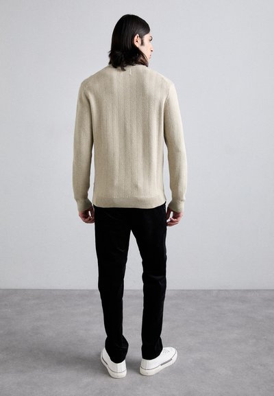 Pull en tricot texturé beige clair, avec un col et des poignets côtelés, associé à un pantalon noir à jambes droites et des baskets blanches.