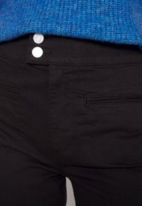 Gros plan sur le devant d'un pantalon noir avec deux boutons argentés et des poches passepoilées, associé à un ourlet de pull en maille bleu visible à la taille.