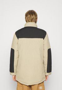 Polstret jakke med beigefarget kropp og sorte skulderpaneler, med høy krage og lange ermer. Stoffet virker glatt og holdbart.