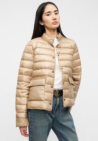 Μπεζ επενδεδυμένο μπουφάν με ψηλό γιακά, διαθέτοντας κούμπωμα με κουμπιά, δύο μπροστινές τσέπες και ομαλή, γυαλιστερή υφή.