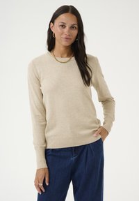 Pull beige à manches longues en matériau doux, avec un col rond et un ourlet côtelé. Porté avec un pantalon en denim bleu ample.