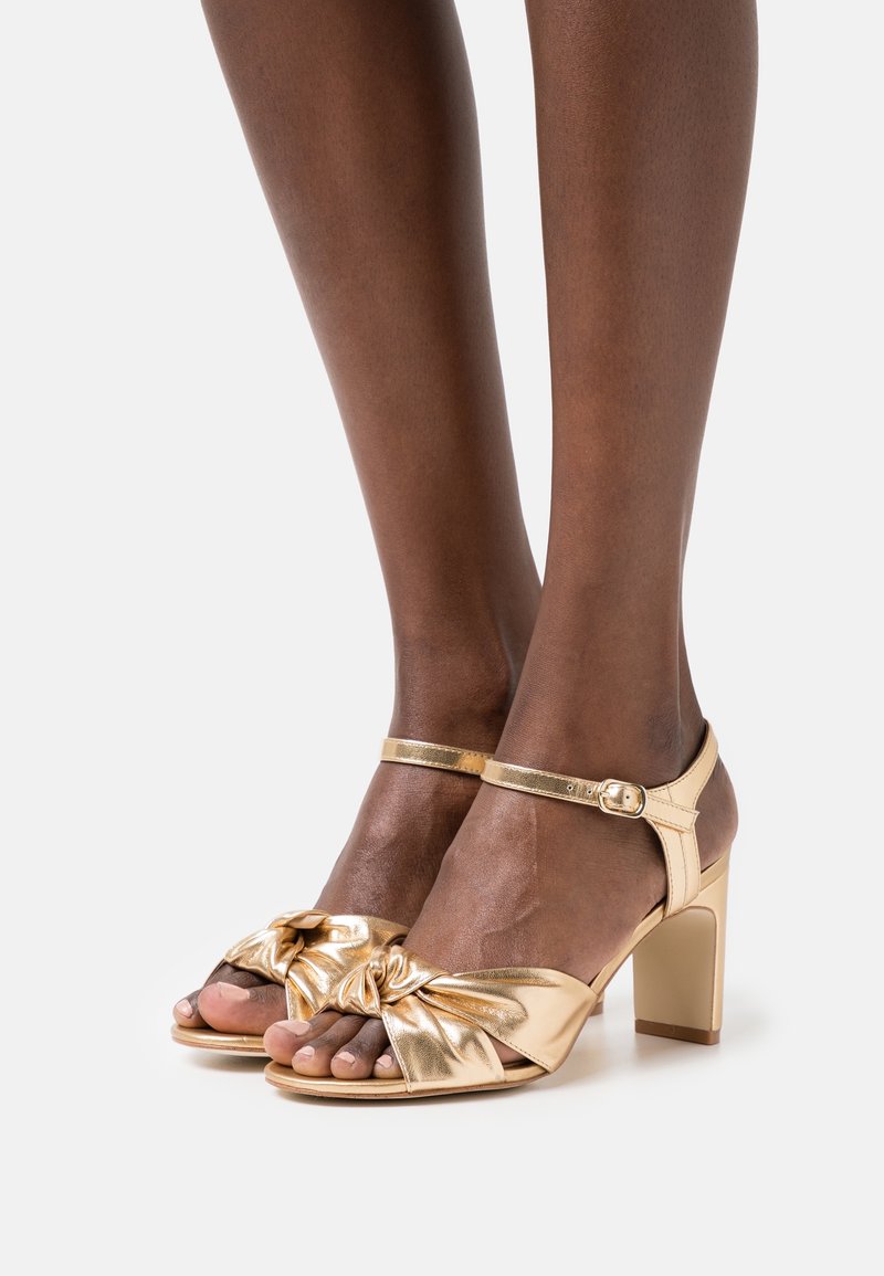 Jambes portant des sandales à talons métalliques dorées à bouts ouverts avec brides à la cheville et détail de nœud froncé sur les brides avant.