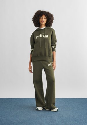 Young woman standing on blue carpet wearing olive green Nike hoodie and matching wide-leg pants with white sneakers.