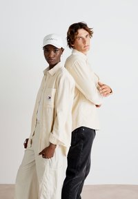 Dos modelos están de espaldas uno al otro. Uno lleva una camisa crema de botones y pantalones color blanco roto; el otro lleva una camisa beige y jeans negros.