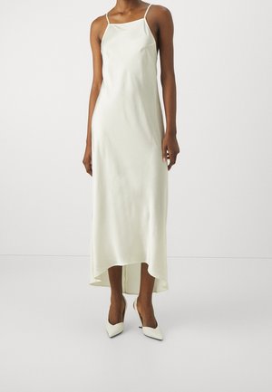 Femme portant une robe nuisette sans manches, écrue, longue jusqu'aux chevilles, avec de fines bretelles et des escarpins à talons pointus, debout sur un fond blanc uni.