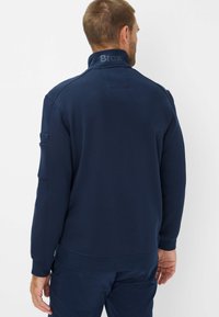 Navy pullover made of soft fabric, featuring a high collar with "Brax" logo, ribbed cuffs, and a side pocket on the left sleeve.