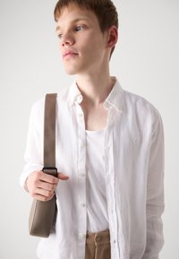 Hvit linlangermet skjorte, beige skulderveske og lys tanktop. Modellen har kort hår og blå øyne, med hender som holder veske strap.
