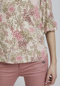 Blumenbluse in Creme mit rosa, grünen und braunen Mustern, locker sitzend und aus strukturiertem Stoff. Kombiniert mit hellrosa, schmal geschnittenen Hosen.