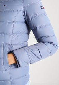 Ljusblå puffjacka med horisontellt quiltad design, utrustad med en ficka och logotypdetalj på ärmen. Mjuk, slät textur.