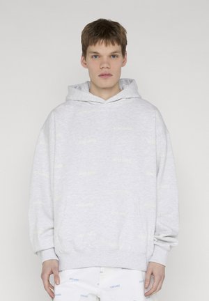 Junger Mann mit kurzen Haaren trägt einen hellgrauen Kapuzenpullover und weiße Hose mit dezenten, wiederholten Markenlogos vor einfachem hellen Hintergrund.