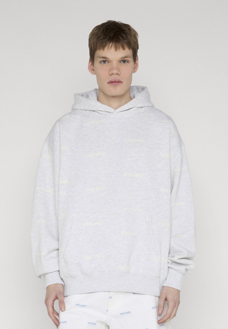 Jeune homme aux cheveux courts portant un sweat à capuche gris pâle et un pantalon blanc avec de faibles logos de marque répétés sur un fond clair uni.