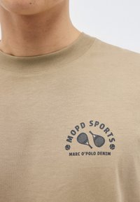 Μπεζ μπλουζάκι με στρογγυλή λαιμόκοψη και σκούρο λογότυπο με το κείμενο "MOPD SPORTS" και "MARC O'POLO DENIM" που απεικονίζει δύο σταυρωτές ρακέτες padel και μπάλες τένις.