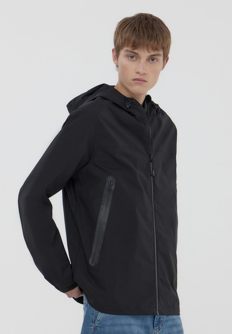 Giacca nera con cappuccio realizzata in materiale leggero, dotata di chiusura a zip frontale, tasca laterale con zip e polsini elasticizzati. Texture liscia.