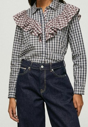 Kvinde iført en ternet bluse med lange ærmer og flæsedetaljer over brystet, kombineret med højtaljede mørkeblå jeans.