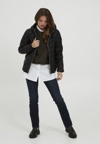Veste matelassée noire avec capuche, dotée d'une fermeture éclair à l'avant et de poches latérales, portée sur un pull vert et une chemise à col blanche, avec un jean sombre.