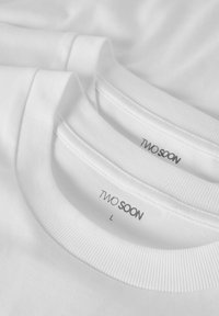 To hvide t-shirts stablet, med ribstrikkede rundhalser. Mærker viser "TWO SOON" og størrelse "L." Glat tekstur uden synlige mønstre.