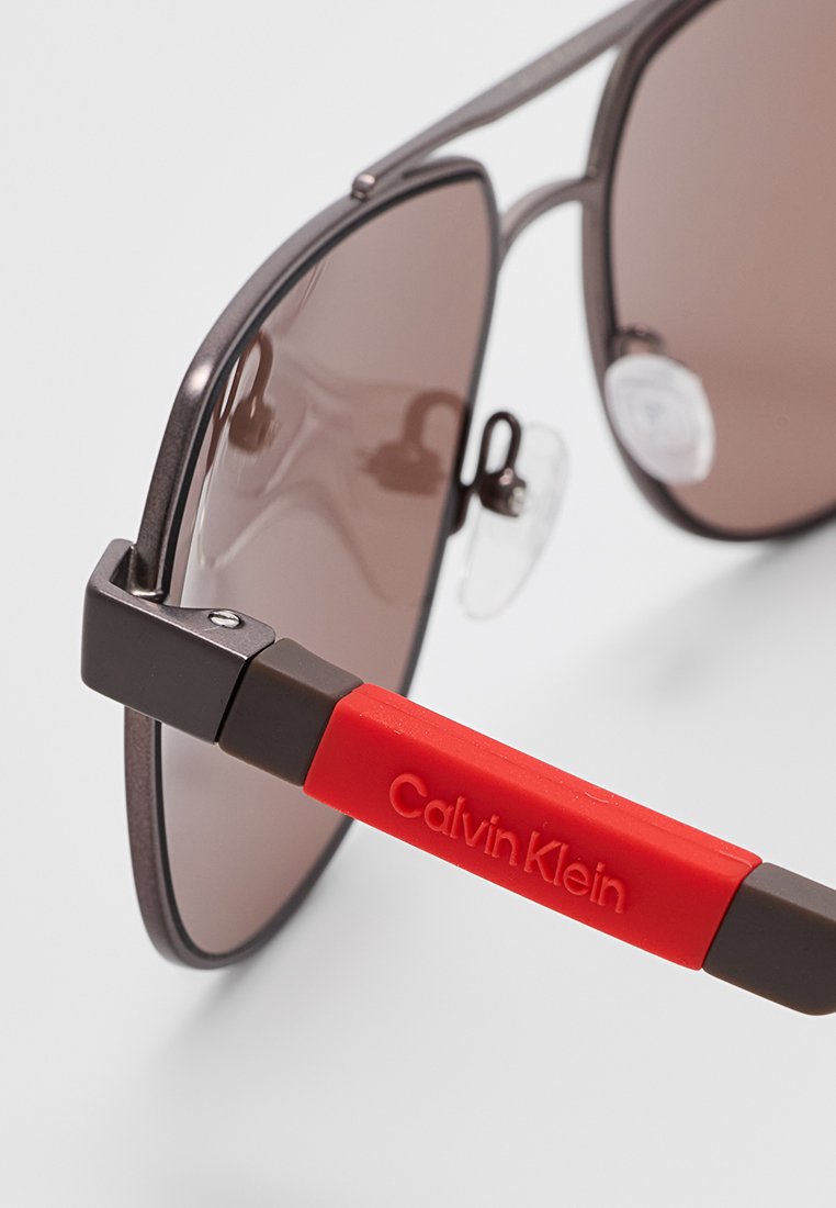 Gafas de sol con lentes marrones, montura gris metálica y patillas de goma roja con el logo en relieve "Calvin Klein" para agarre y estilo.