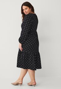 Vestido negro con lunares, con dobladillo en capas, mangas largas abullonadas y un torso ajustado. Combinado con zapatos de color natural, que muestran una textura suave.