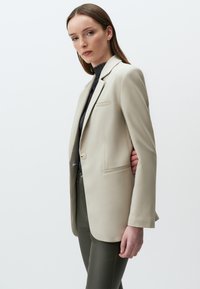 Beige maßgeschneiderter Blazer mit einem einzigen Knopf, Reverskragen und Seitentaschen, kombiniert mit eng anliegenden olivgrünen Hosen. Glatte Textur, strukturiertes Design.