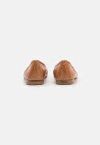 Un par de mocasines de cuero marrón mostrados desde atrás sobre un fondo blanco con tacones de madera apilados bajos.