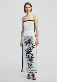 Maxikleid aus weißem Stoff mit einem schwarzen tropischen Pflanzenmuster, dünnen verstellbaren Trägern und seitlichen Schlitzen. Schwarze Akzente entlang des Ausschnitts und der Seiten.