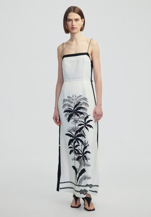 Maxi dress in white fabric featuring a black tropical plant print, thin adjustable straps, and side slits. Black accents along the neckline and sides.