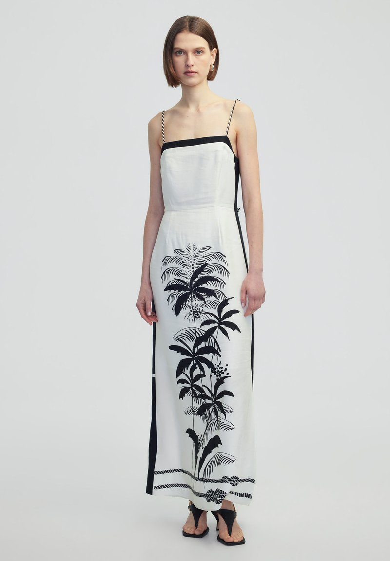 Robe maxi en tissu blanc présentant un imprimé de plantes tropicales noires, avec des bretelles fines et réglables, ainsi que des fentes sur les côtés. Accents noirs le long de l'encolure et des côtés.