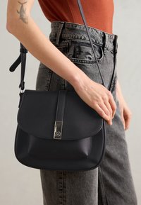 Sac bandoulière en cuir noir avec une fermeture à rabat, quincaillerie en argent et une sangle adjustable. Surface texturée avec des bords arrondis.