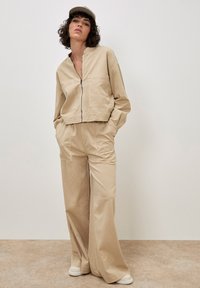 Vrouw in een beige ritsjack en wijde broek met handen in de zakken, die een bijpassende beige pet en witte sneakers draagt.