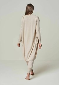 Langer beiger Cardigan mit lässiger Passform, seitlichen Taschen und langen Ärmeln. Über passenden beigen, figurbetonten Hosen getragen. Glatte Textur.