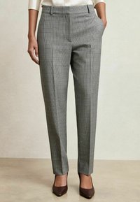 Mujer vistiendo pantalones de cuadros grises ajustados con bolsillos laterales y tacones marrones de punta, de pie sobre un suelo beige.