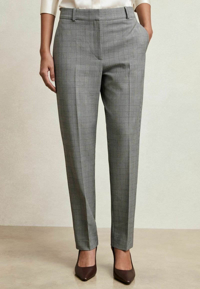 Mujer vistiendo pantalones de cuadros grises ajustados con bolsillos laterales y tacones marrones de punta, de pie sobre un suelo beige.