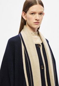 Μπλε ναυτικό πόλο από μάλλινο μείγμα με κρεμ κάθετες ρίγες, διαθέτει στρογγυλή λαιμόκοψη, κούμπωμα με κουμπιά και απαλή κασκόλ. Λεία υφή με φυσική κίνηση.