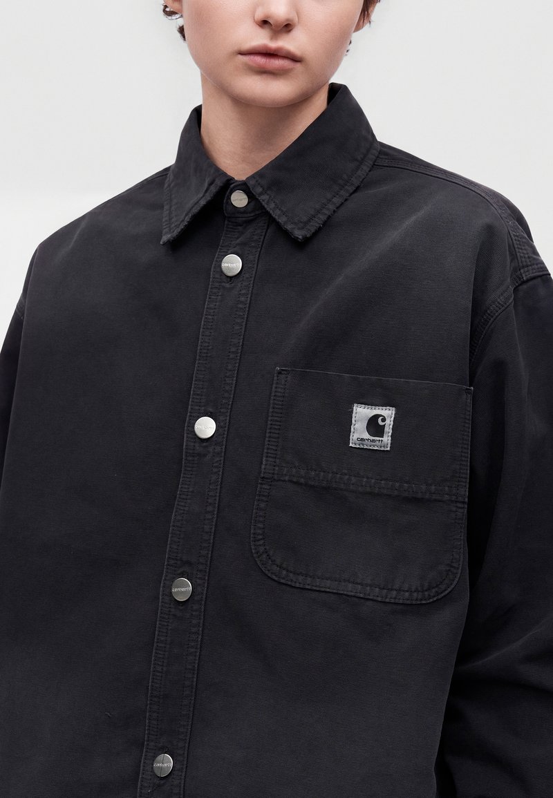 Junge Person trägt ein schwarzes Carhartt-Hemd mit Knöpfen, einer Brusttasche und sichtbarem Markenlogo auf der Tasche.