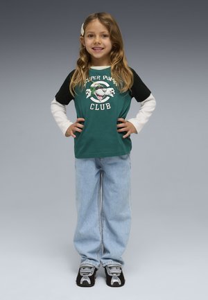 Ragazza giovane con capelli lunghi in piedi sorridente, mani sui fianchi, indossa una maglietta verde e nera "Super Puma Club", jeans chiari e scarpe da ginnastica nere.