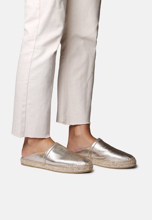 Nohy v metalických slip-on espadrilloch v štýle mules, skombinované s krémovými skrátenými nohavicami na neutrálnej pozadí.