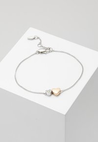 Bracelet chaîne en argent avec deux breloques en forme de cœur : une en or, une en argent ornée de petits cristaux intégrés, présenté sur un bloc blanc.