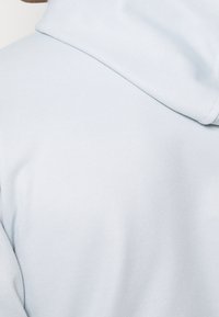Svetle modrá mikina s kapucí vyrobená z texturované tkaniny, s měkkou kapucí a volným střihem, která zdůrazňuje hladké švy a jednoduchý design.