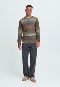 Suéter de punto para hombres en tonos degradados de verde y marrón, con cuello redondo y puños acanalados, combinado con pantalones rectos de pierna oscura y zapatillas grises.